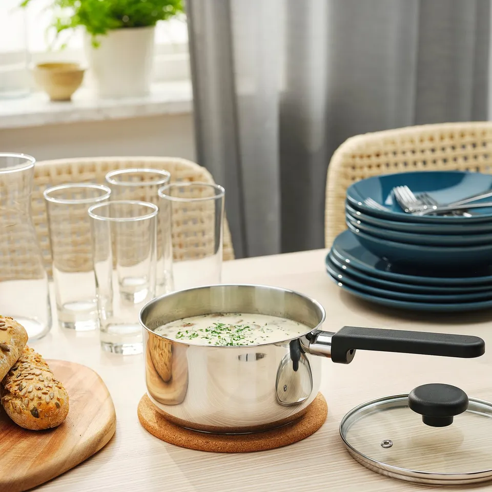 Stainless steel saucepan with black handle and lid sits on wooden table, filled with creamy soup, surrounded by glasses and plates, ready for serving.
