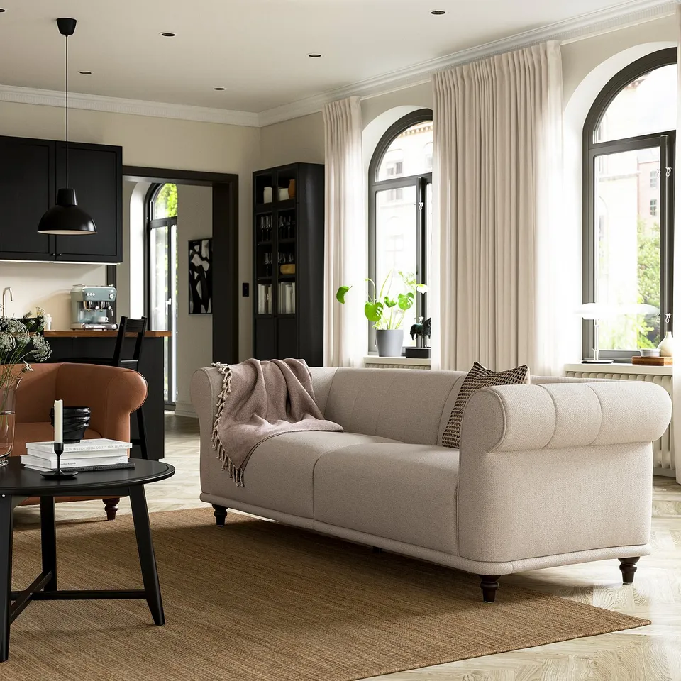 Modern living room with beige sofa, orange chair, black side table, large windows, plant, and kitchen with black cabinets and white counters.