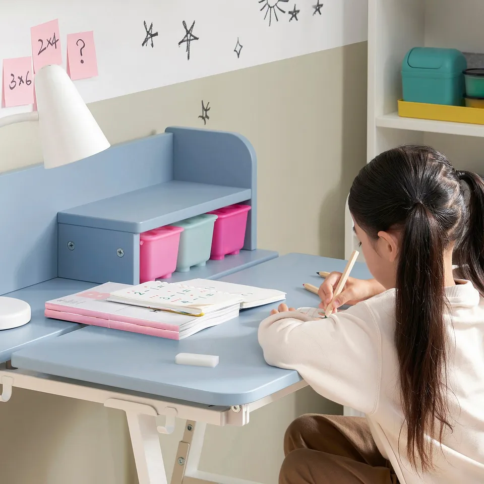 A girl sits at a PIPLÄRKA kids desk, tilting the surface for better posture. The person writes in a workbook beside colourful storage bins and a lamp.
