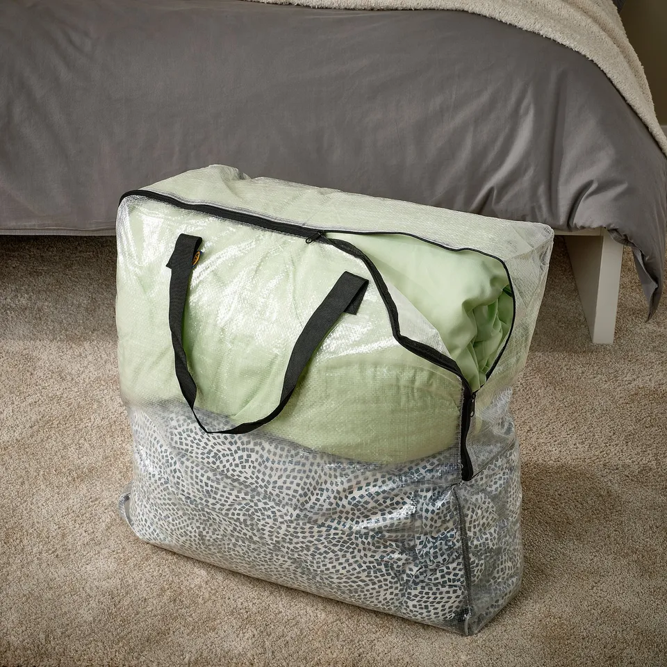 Transparent storage bag with handles on carpet next to bed, folds flat when empty.