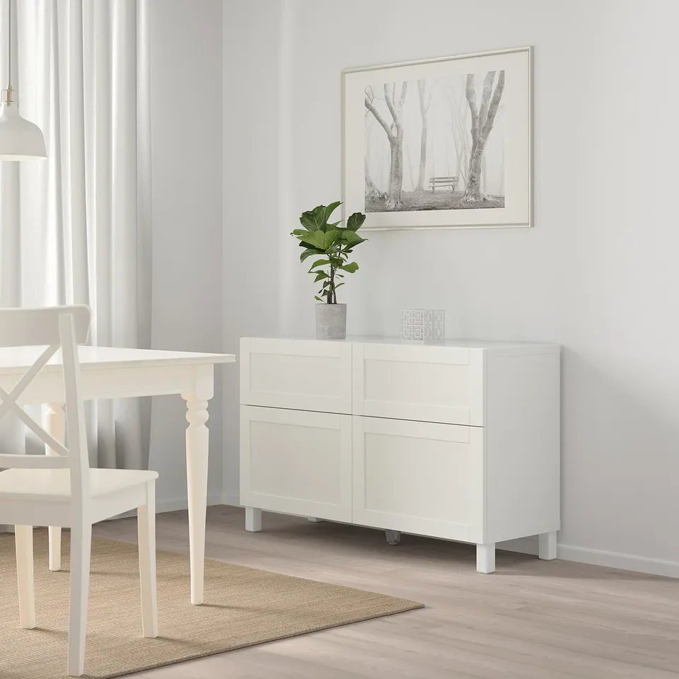 Modern white BESTÅ storage unit, narrow and tall, beside dining table. Decorated with plant and framed artwork.