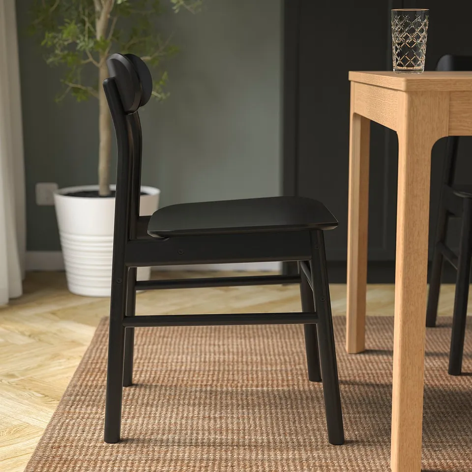 Black modern chair with wooden frame and seat, placed near wooden table and plant.