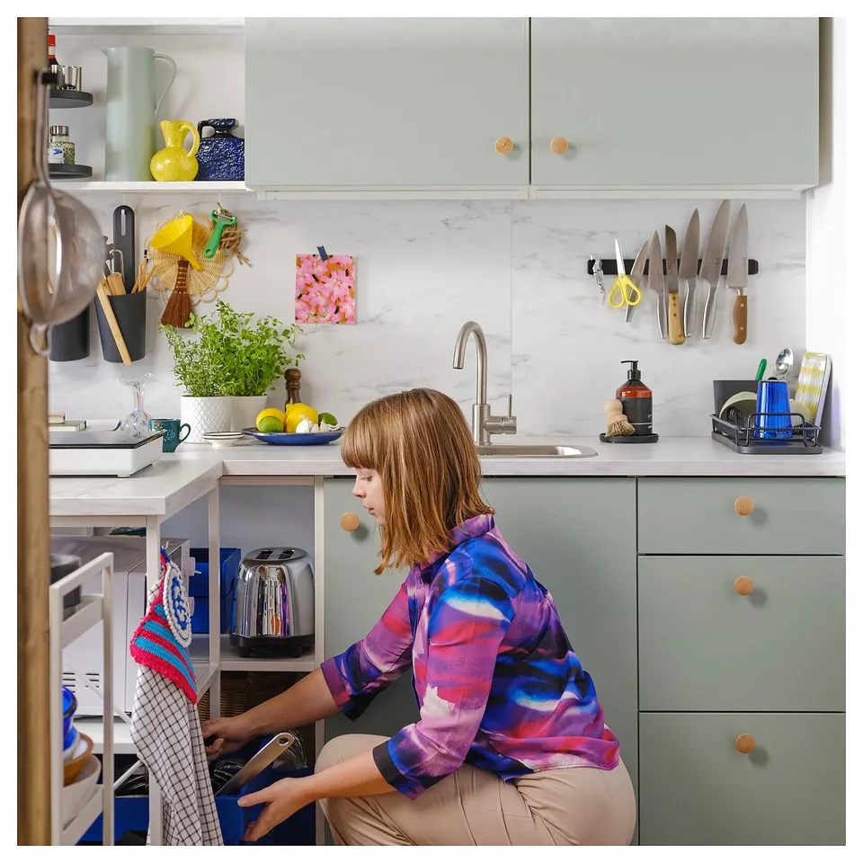 Person kneeling by green oven with knobs, plants, utensils.