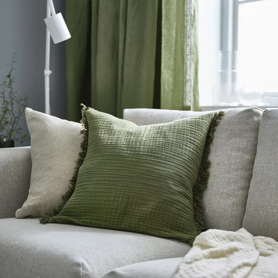 Green VALLKRASSING cushion on grey sofa, fringed, surrounded by pillows. Green curtains, lamp in background.