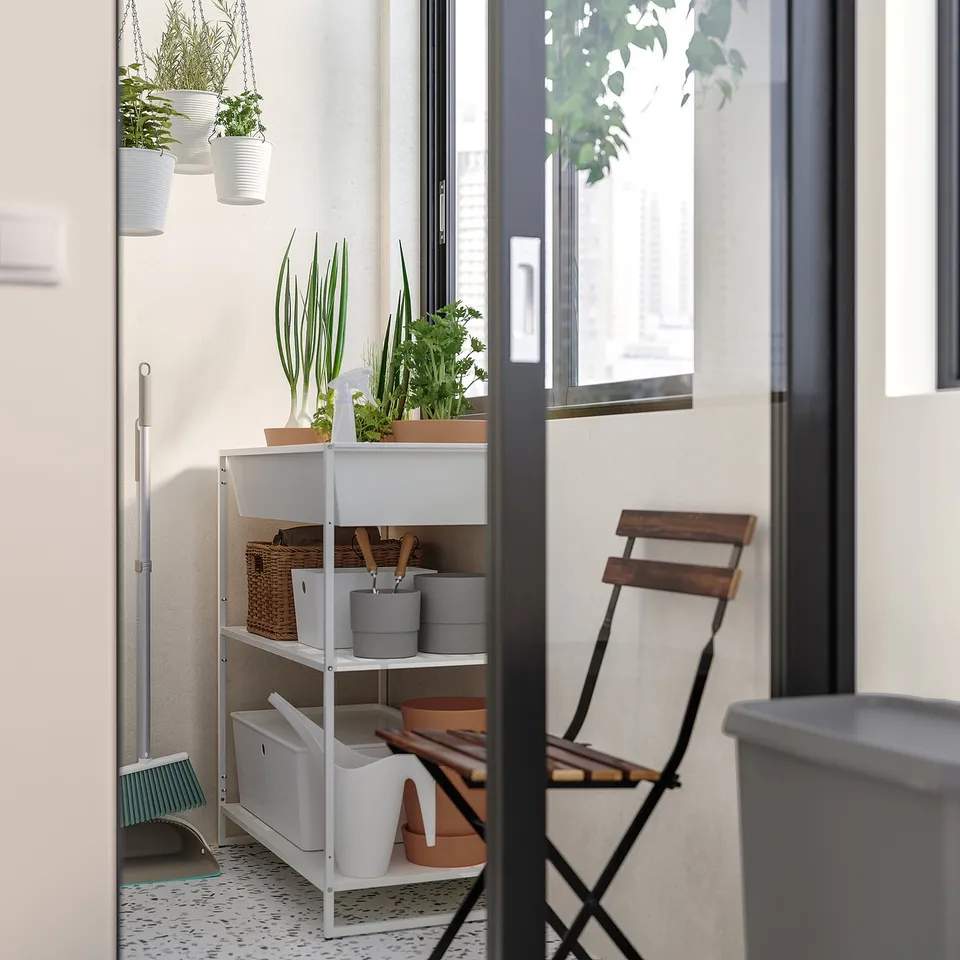 White metal shelving unit with potted plants, wicker baskets, and cleaning tools in a bright room with a wooden chair.