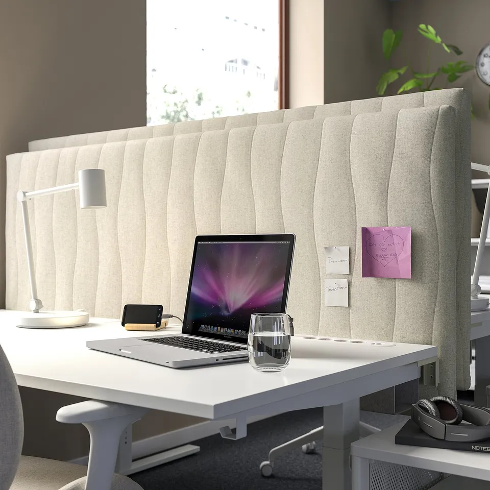 Acoustic screen divides workspace. Dense fabric, wooden core. Desktop setup with laptop, glass.