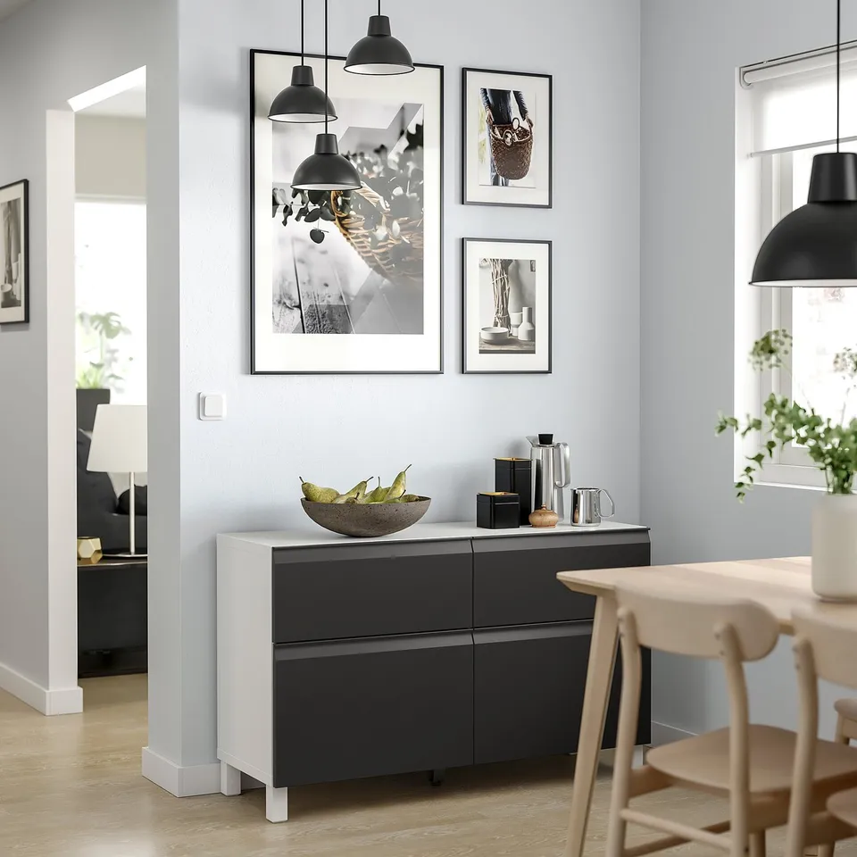 Modern dining area with a white and black BESTÅ sideboard, light wooden chairs, hanging lamps, and framed photos on light grey walls.