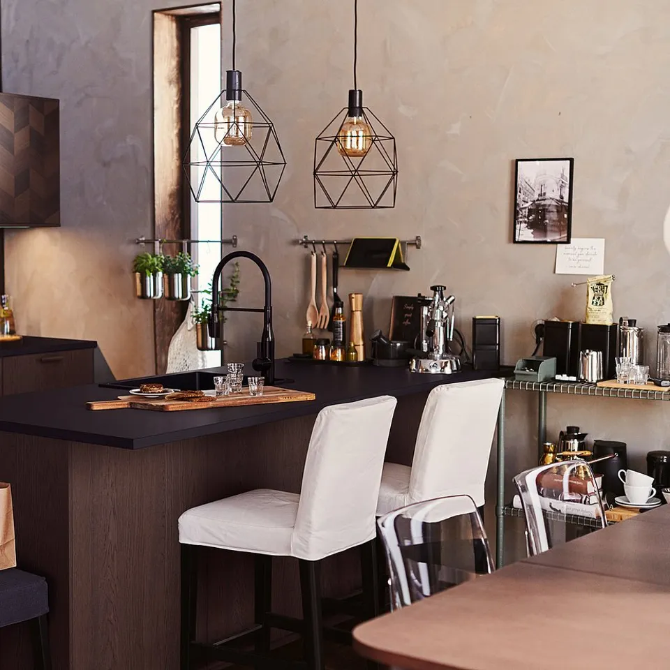 Modern kitchen with dark wooden cabinets, a countertop with two stools, hanging black pendant lamps, and various kitchenware.