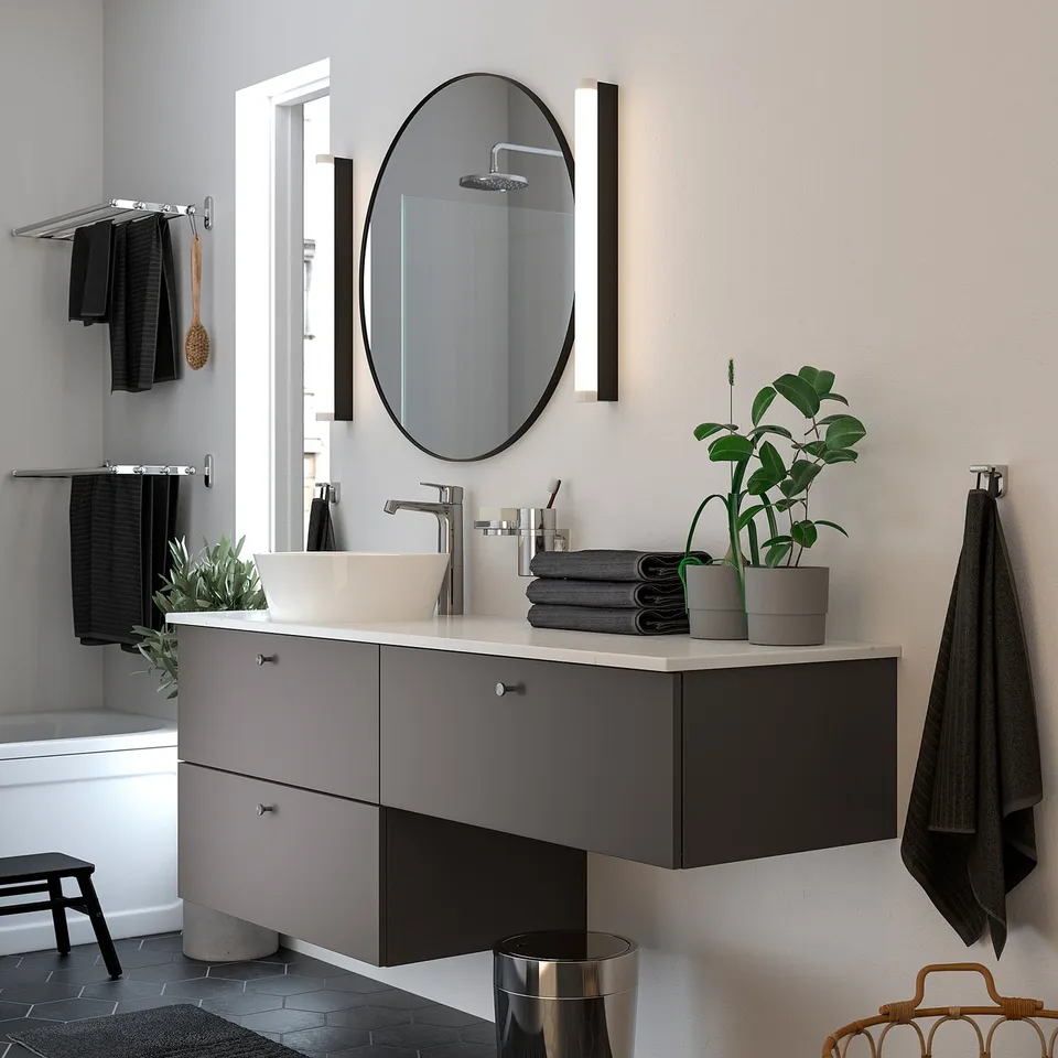 Modern bathroom with LINDBYN round black-framed mirror above sleek grey vanity, plants, and black towels.