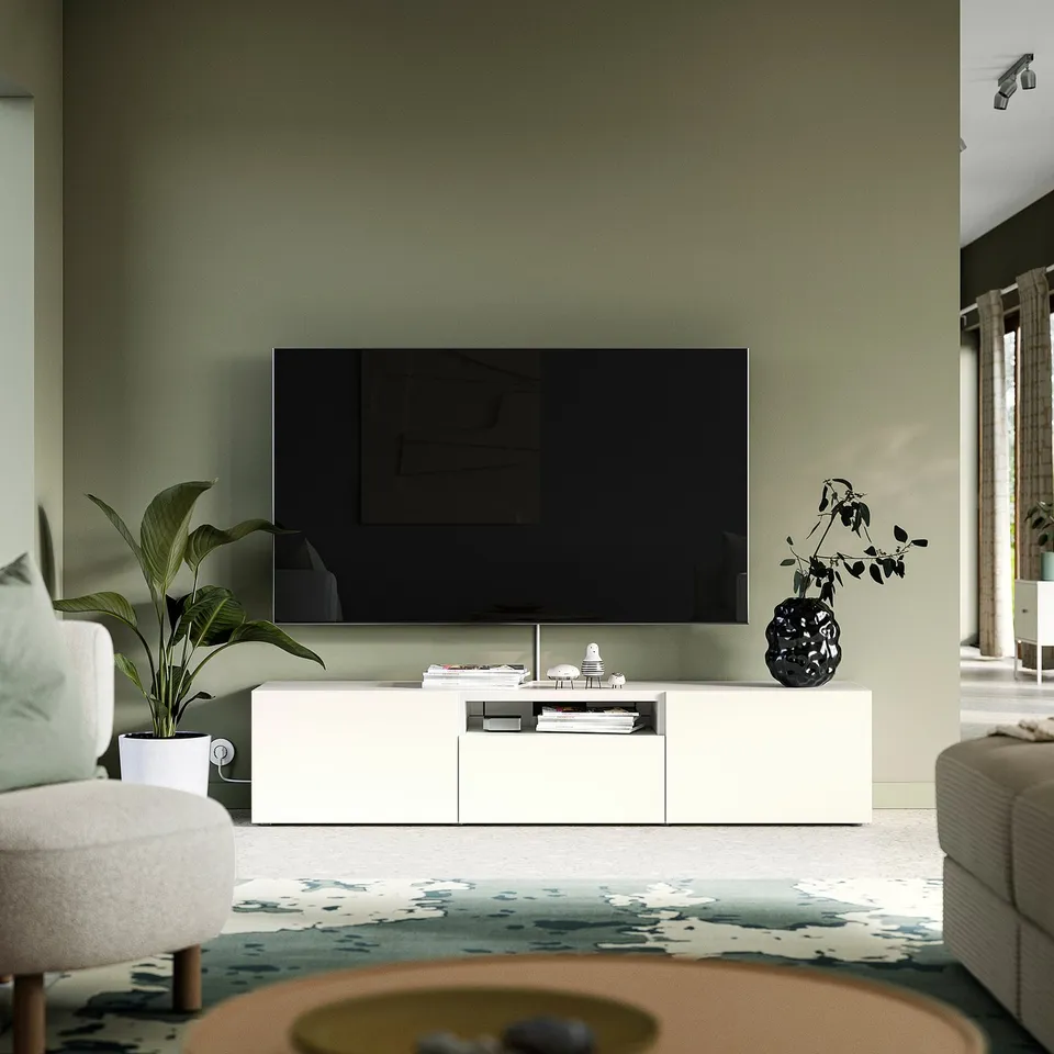 Modern living room with BESTÅ tv stand, flat-screen tv, books, and decor. White armchair left, beige sofa right, grey rug, sage wall, natural light.