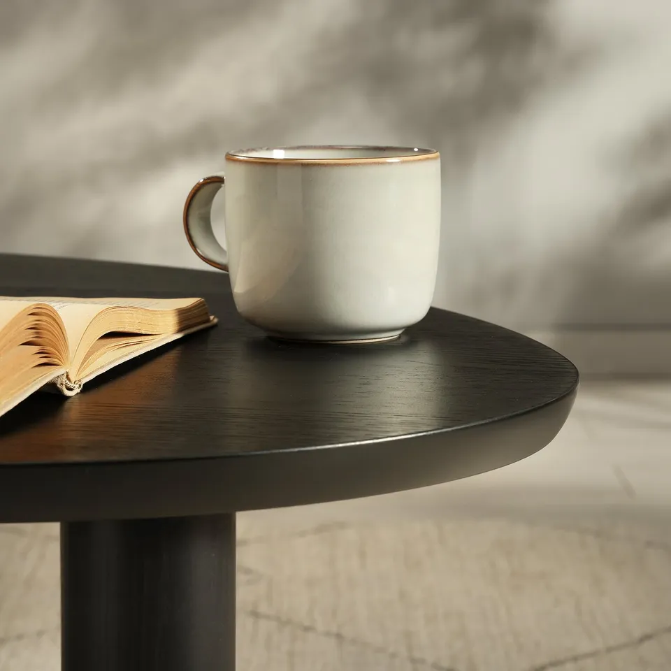 Ceramic mug on sleek, dark round table beside open book, showcasing quality wood texture and sophisticated design.