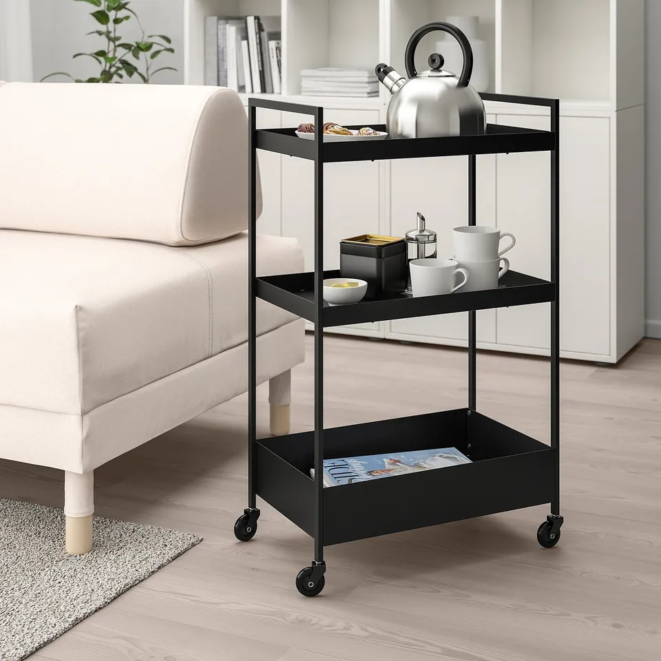 Black 3-tier ikea nissafor trolley beside white couch, holding tea set. Compact & mobile storage.