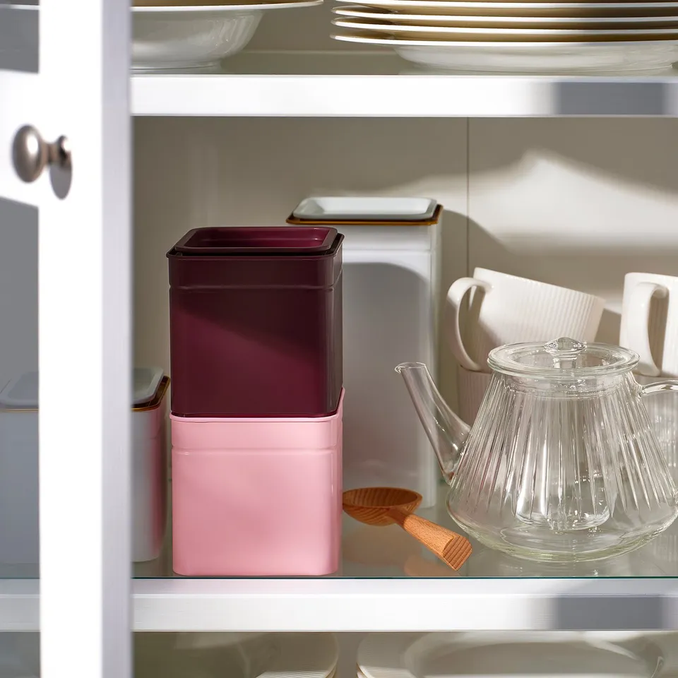 A kitchen shelf with a pink and brown metal tin for storing tea or coffee, a clear glass teapot, and white mugs.