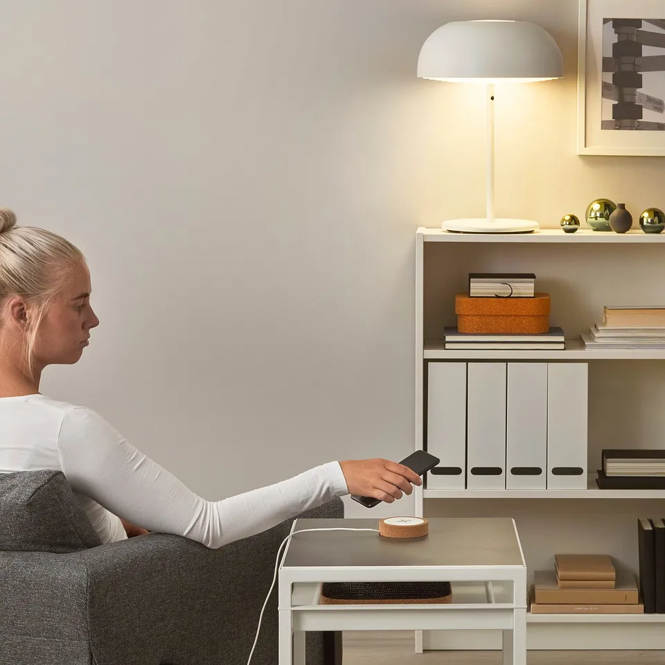 A person uses a NORDMÄRKE wireless phone charger on a table, lighting a lamp nearby, creating a cosy, minimalist workspace.