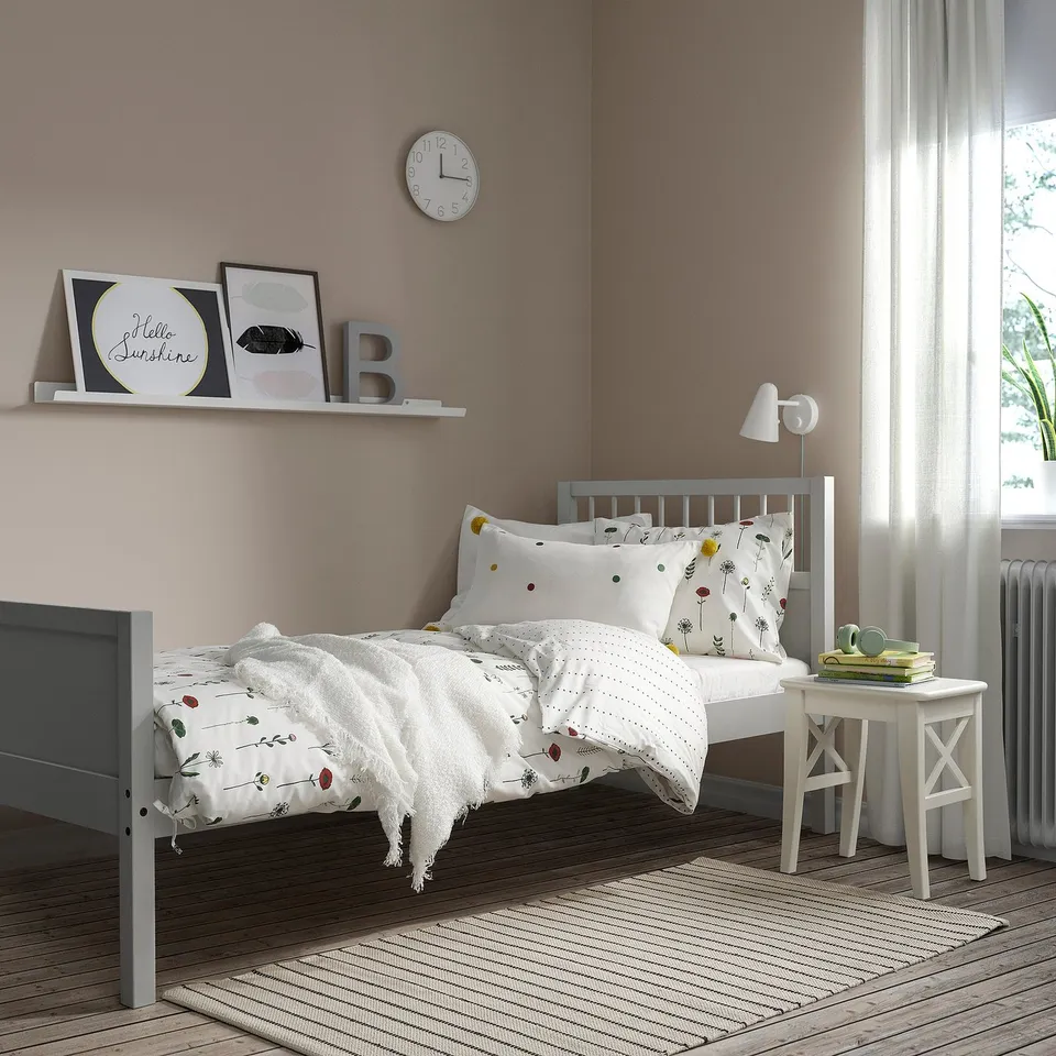 Small grey SMYGA bed in cosy room, floral bedding, white bedside table.