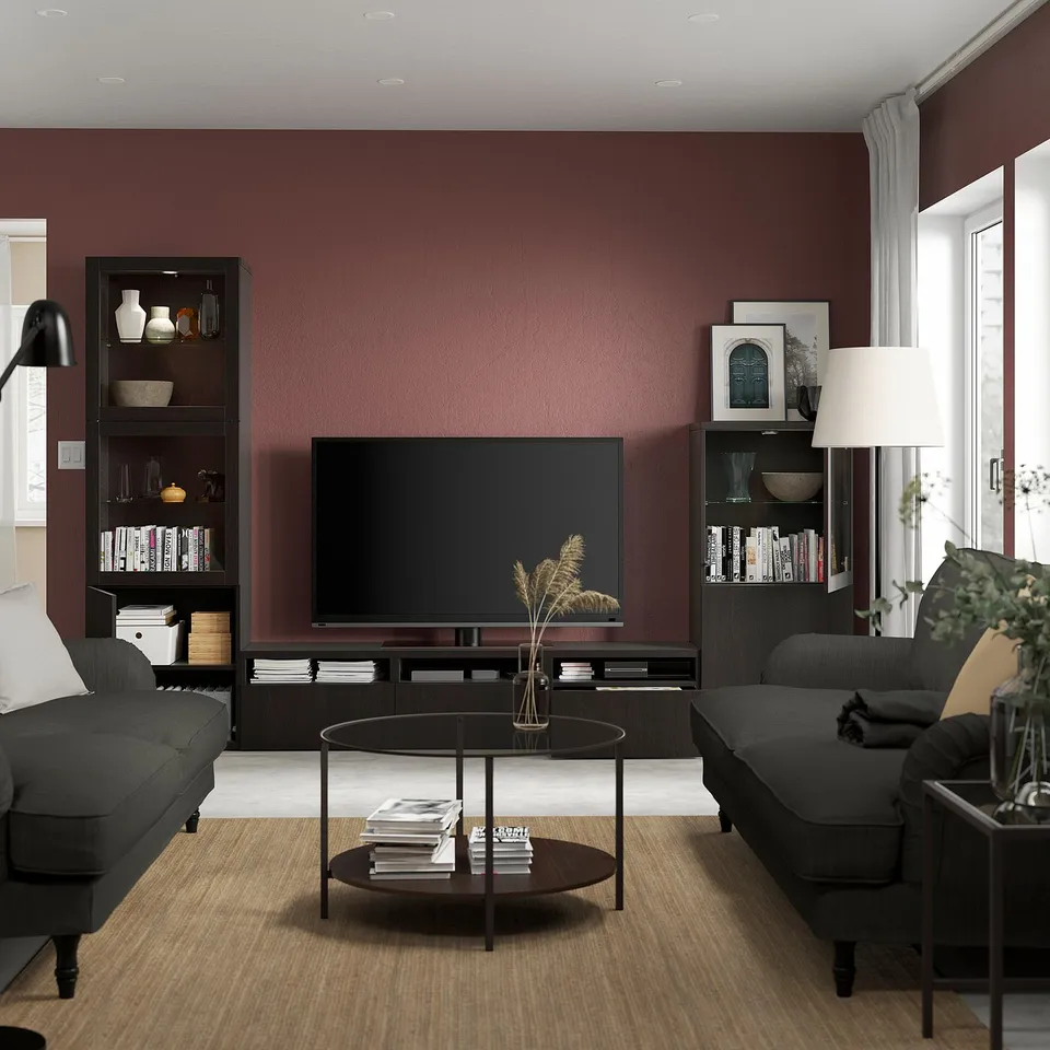 Modern living room with red walls, grey couches, BESTÅ tv unit, books, decor, flat-screen tv, coffee table, floor lamp, large window.