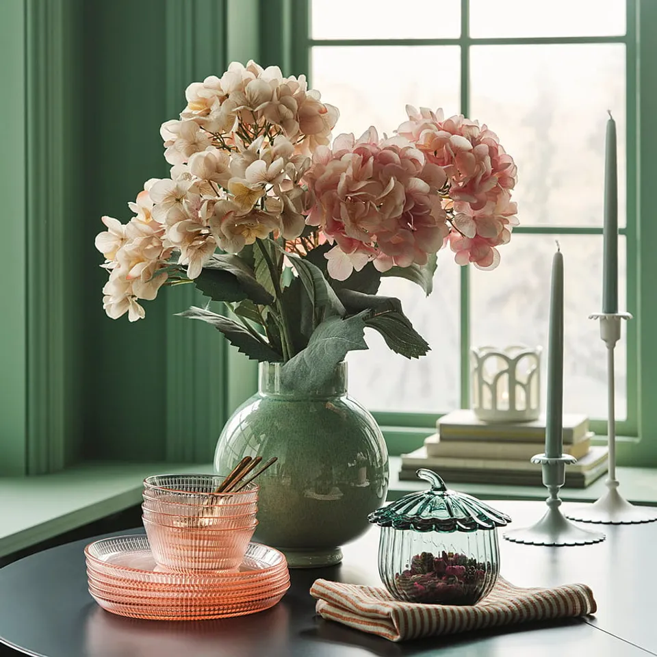 A small green vase with pink & white hydrangeas sits on a table near orange bowls and a jar with dried flowers, candles, and books.