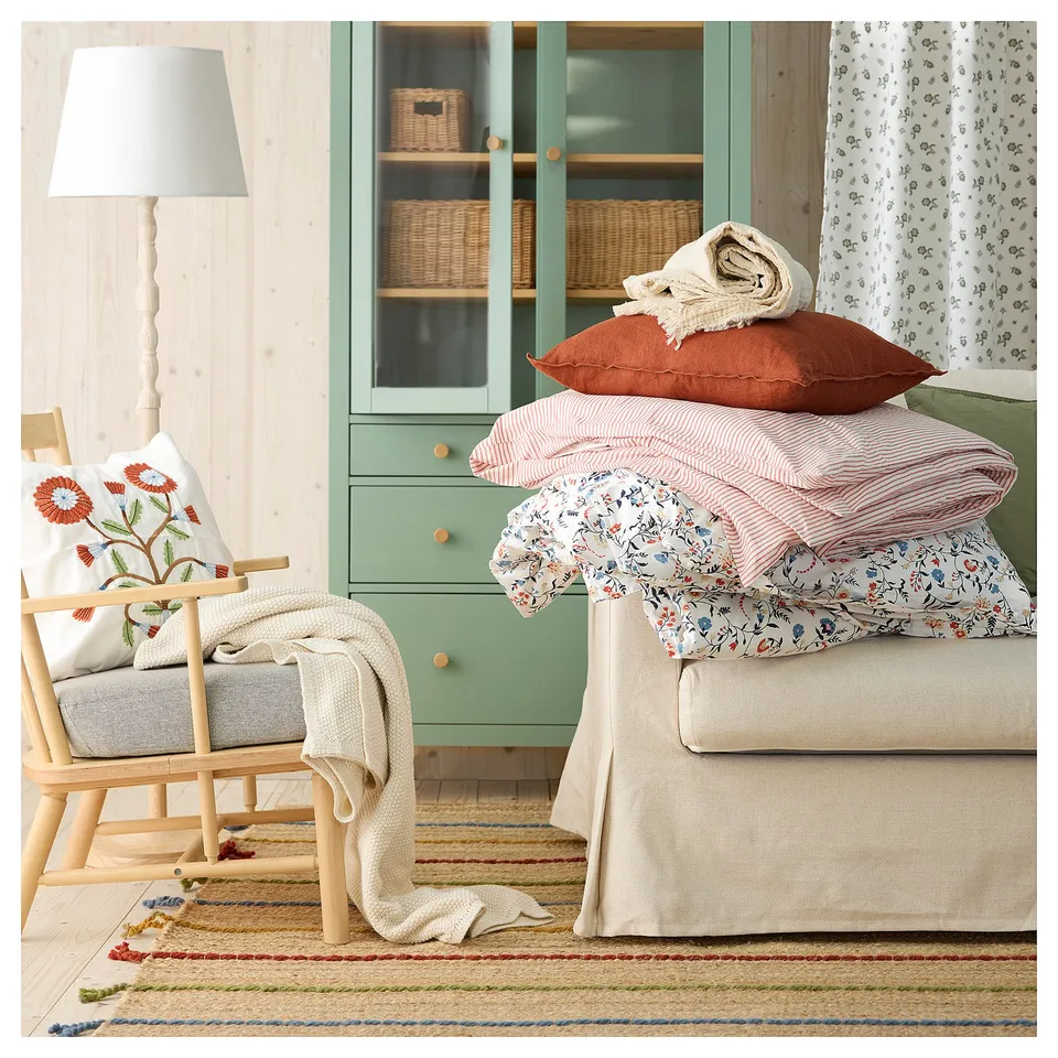 Cosy bedroom with green cabinet, white bed with red and floral pillows, wooden chair, lamp, woven rug.
