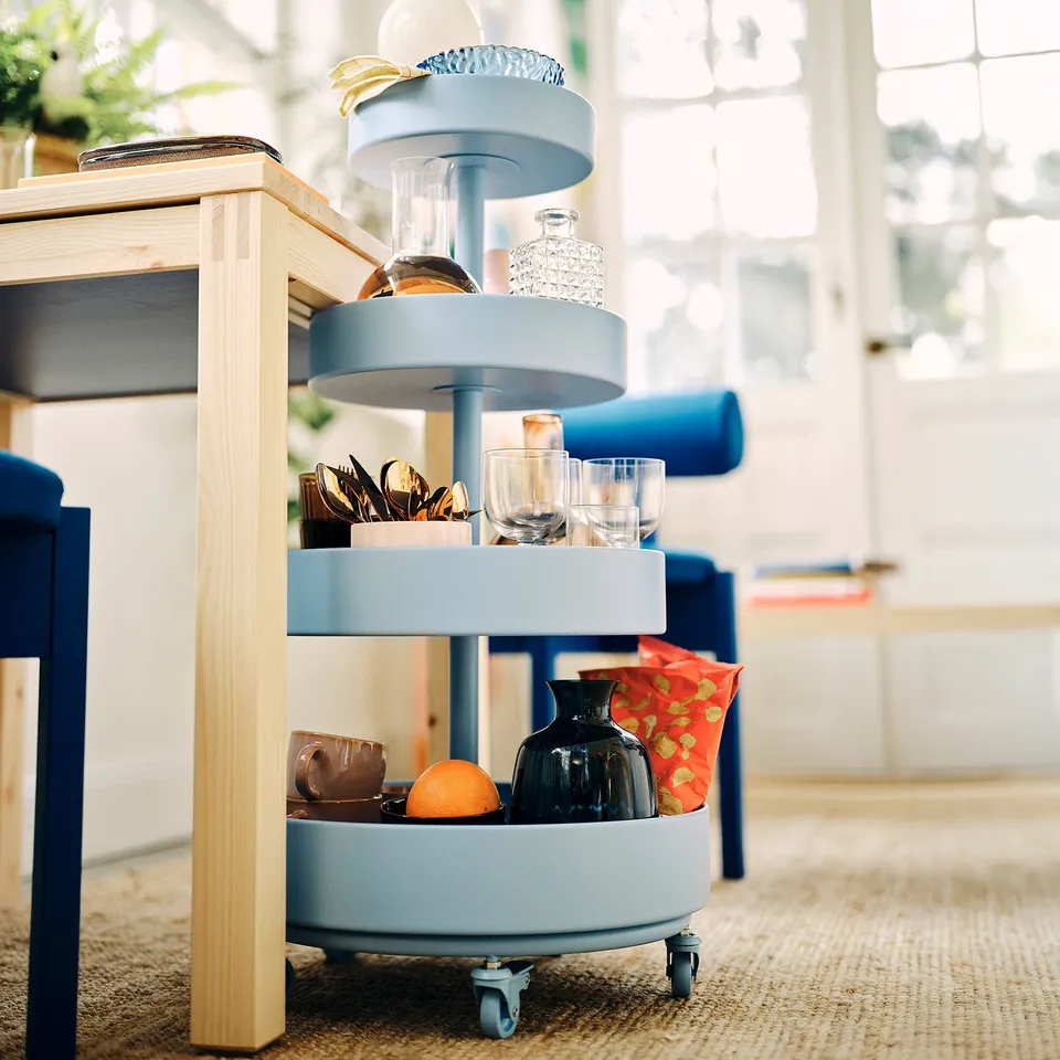 Blue IKEA PS 2026 trolley with rounded shelves, holding glasses, vase, and orange. Tall, sturdy metal, fits neatly under wooden table.