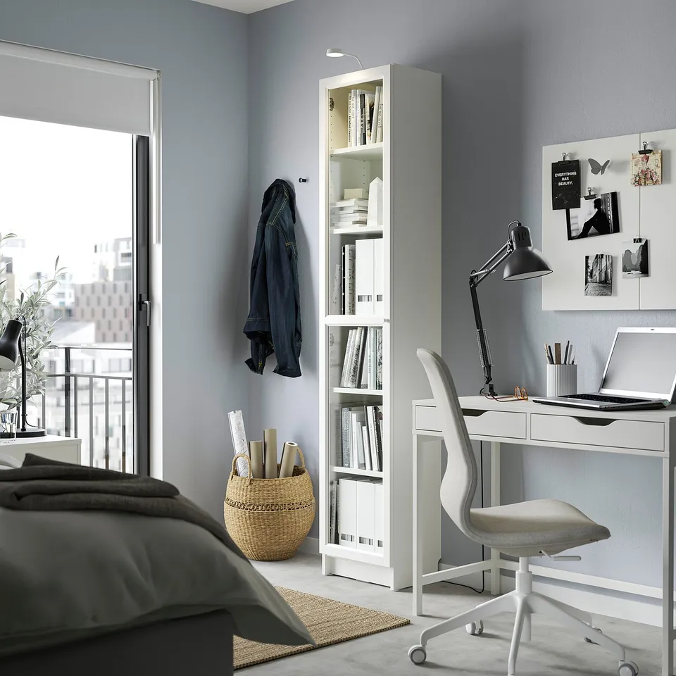 Modern home office with BILLY white bookshelf, desk, & chair. Grey walls, blue jacket, and city view.