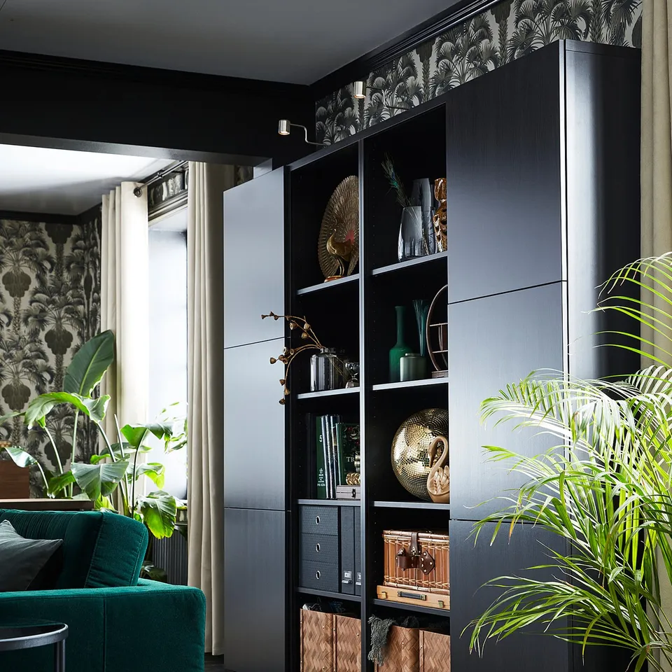 Grey BESTÅ shelves with books and decor, flanked by a green sofa and plants against patterned wallpaper.