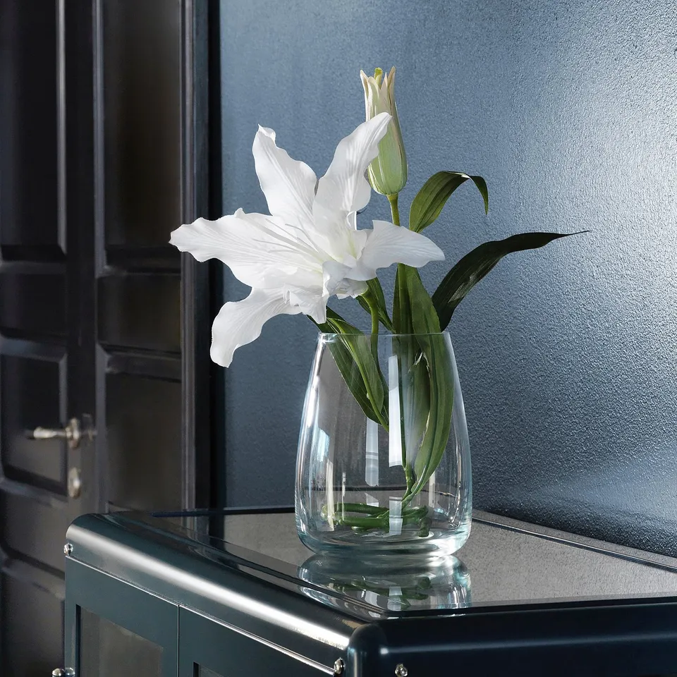 A clear glass vase holding two white lilies on a black surface. The vase is sleek and simple, enhancing the beauty of the flowers.