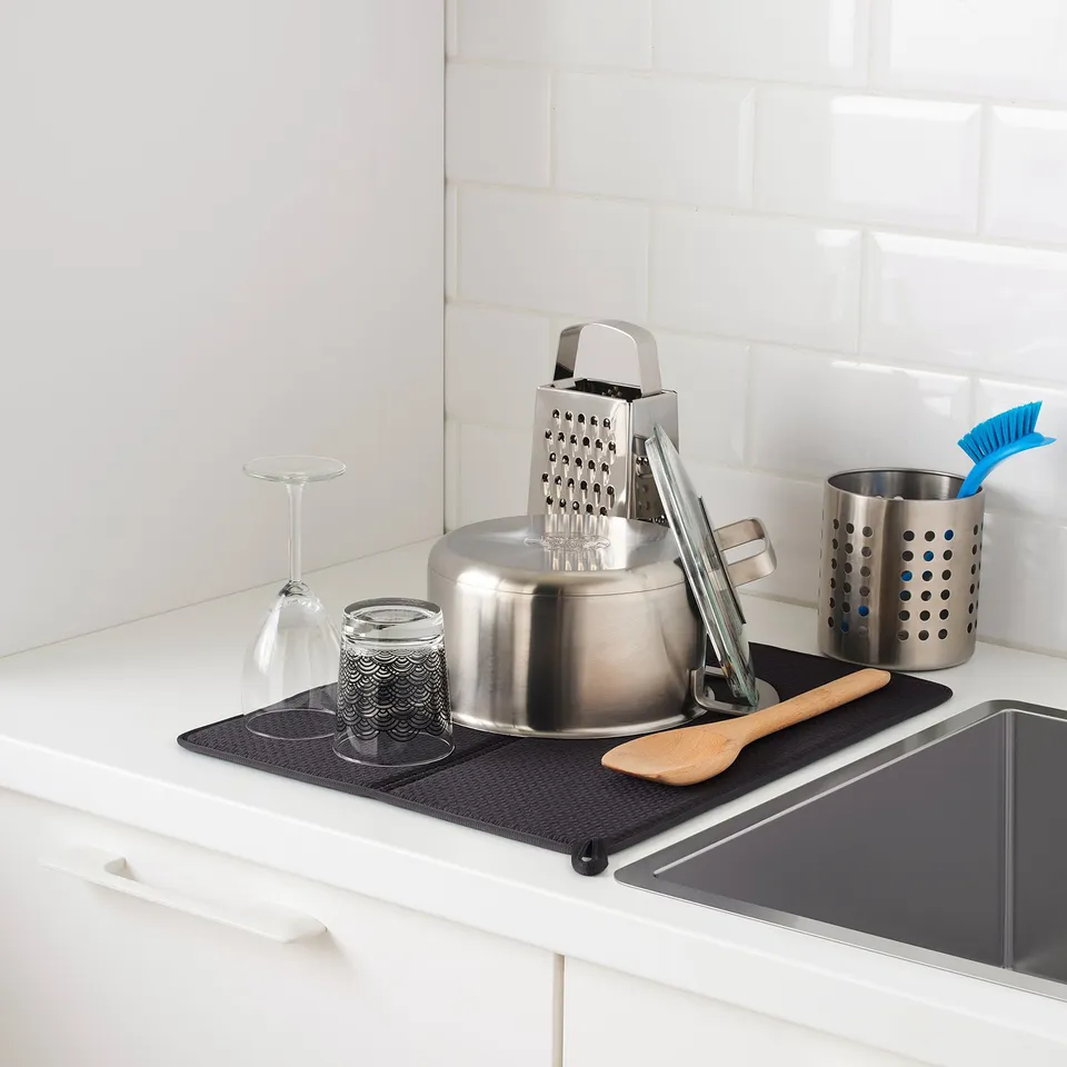 Kitchen sink area with ORDNING stainless steel stand holding a grater, jar, wine glass, and utensil holder with brush, organised on a dark grey mat.
