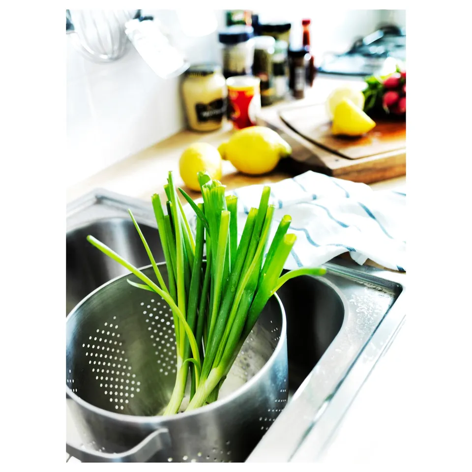 Stainless steel kitchen sink with green onions in strainer. Lemons and cooking utensils nearby.
