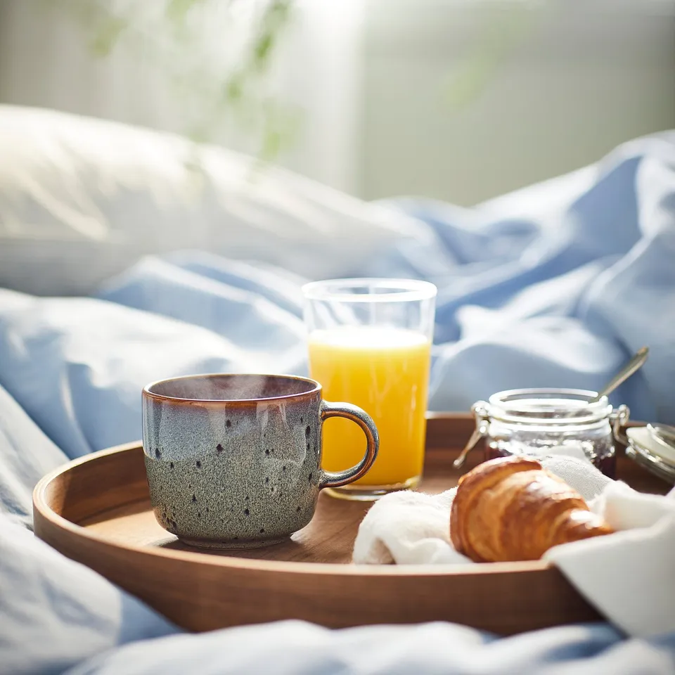 Breakfast tray: blue glazed cup, glass of orange juice, pastry, jam on bed. Mug larger than glass.