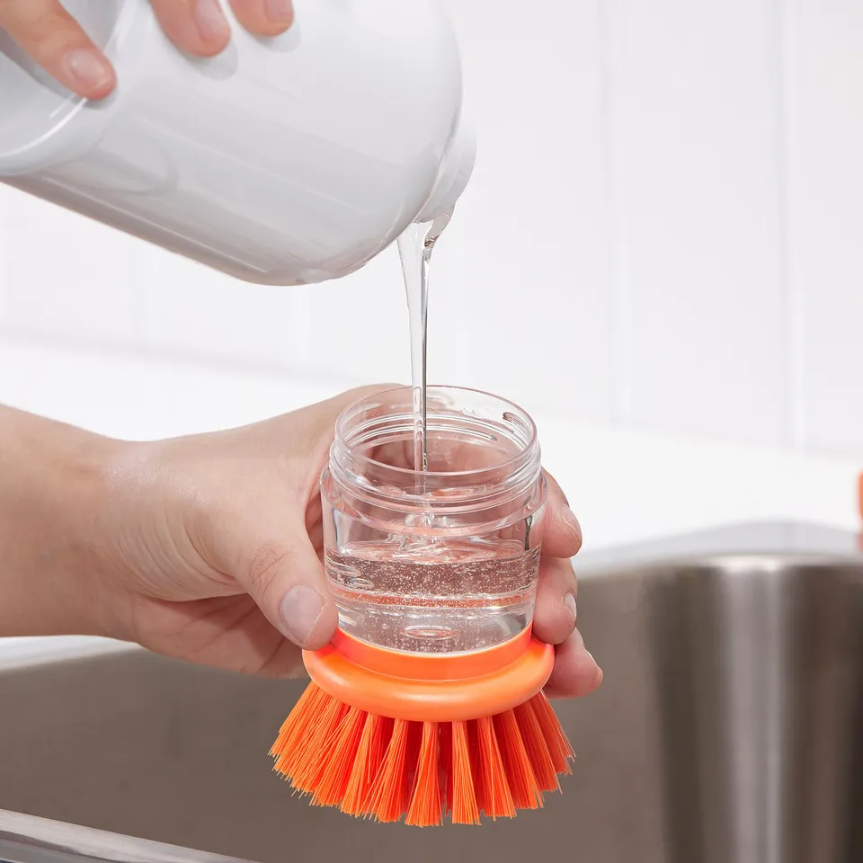 A hand pours liquid from a white container into a small clear jar with an orange brush attached.