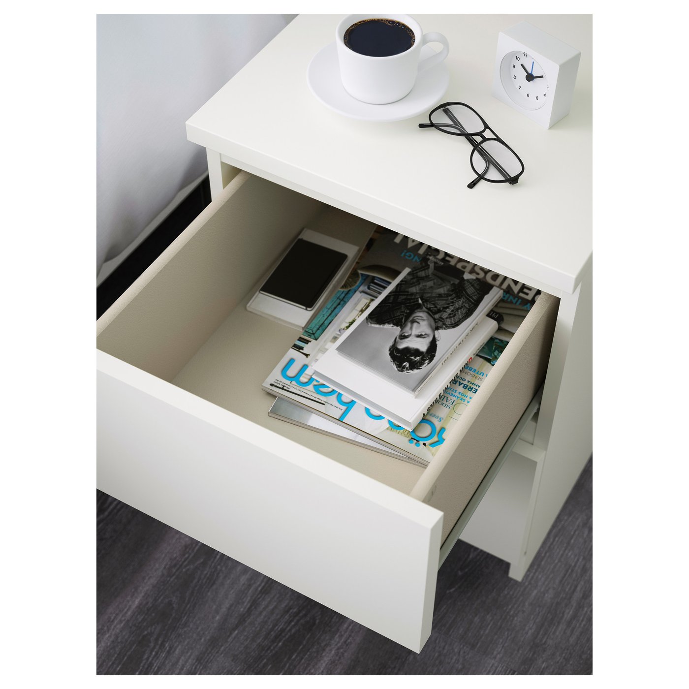 White bedside drawer open, showing magazines, phone, and glasses on top, coffee cup, and clock.
