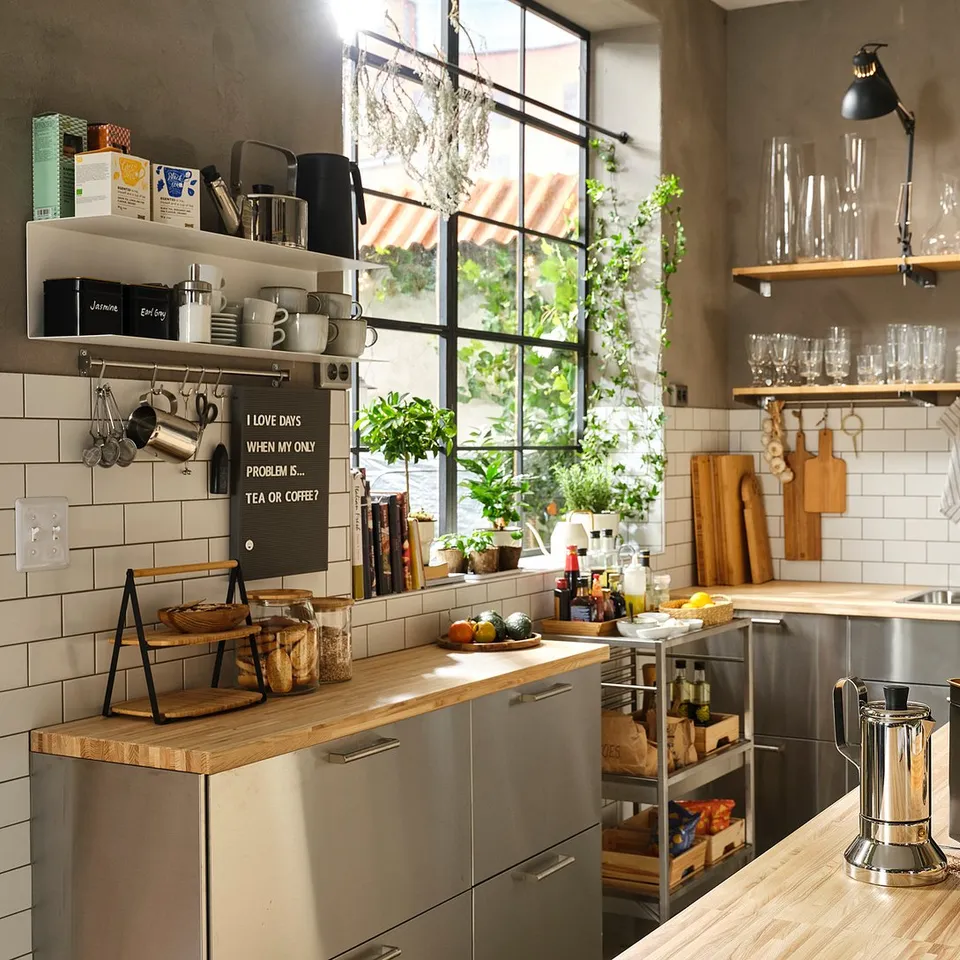Modern industrial kitchen with stainless steel cabinets, wooden countertops, hanging lights, and open shelves displaying dishes and plants.