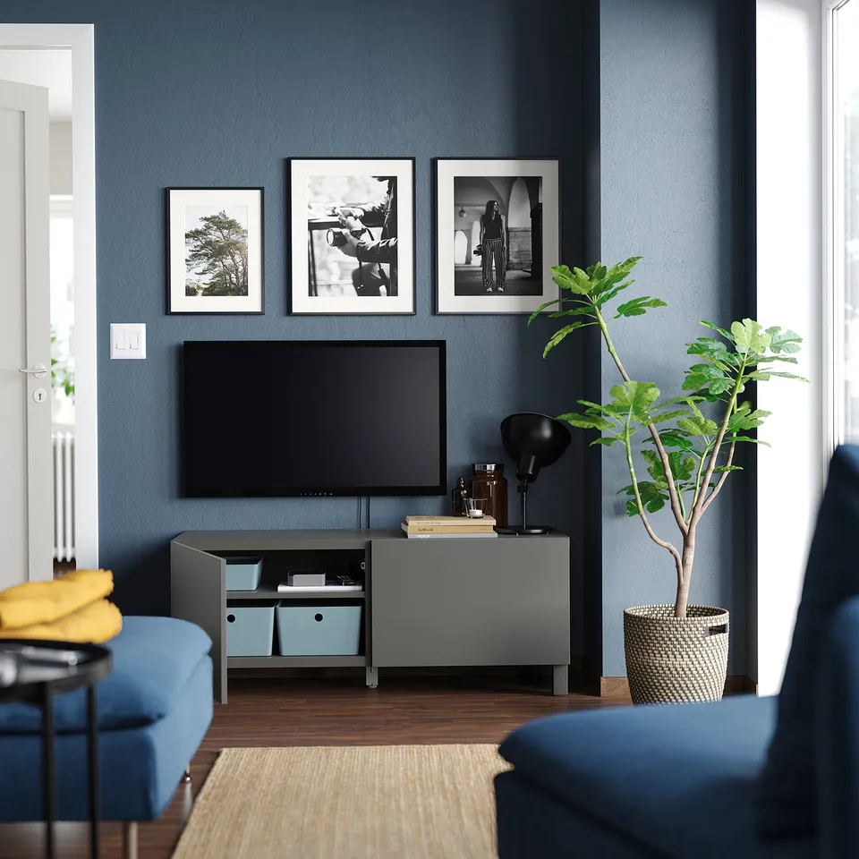 Modern living room with dark blue walls, a grey BESTÅ tv bench, flat-screen tv, blue cushioned chairs, and decorative plants.