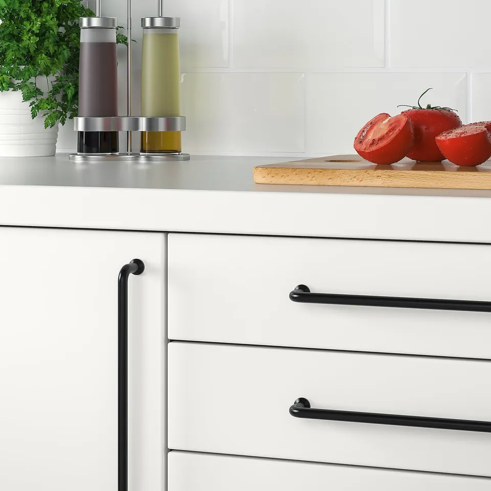 White kitchen drawers with black BAGGANÄS handles; tomatoes on counter, herbs in vase, oil dispensers beside.