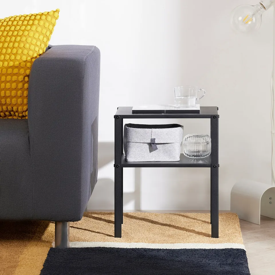 Small black KNARREVIK bedside table next to a grey chair. Holds a glass cup, book, and storage box, ideal for small spaces.