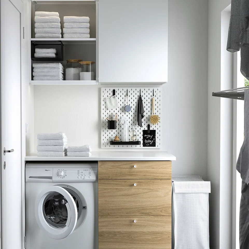 SKÅDIS pegboard with accessories above washing machine, holding towels and small items, surrounded by cabinets and stacks of towels.