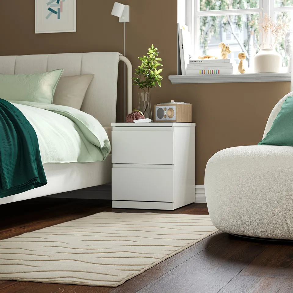 Modern brown bedroom with white STORKLINTA bedside table featuring 2 drawers, positioned between bed and window.