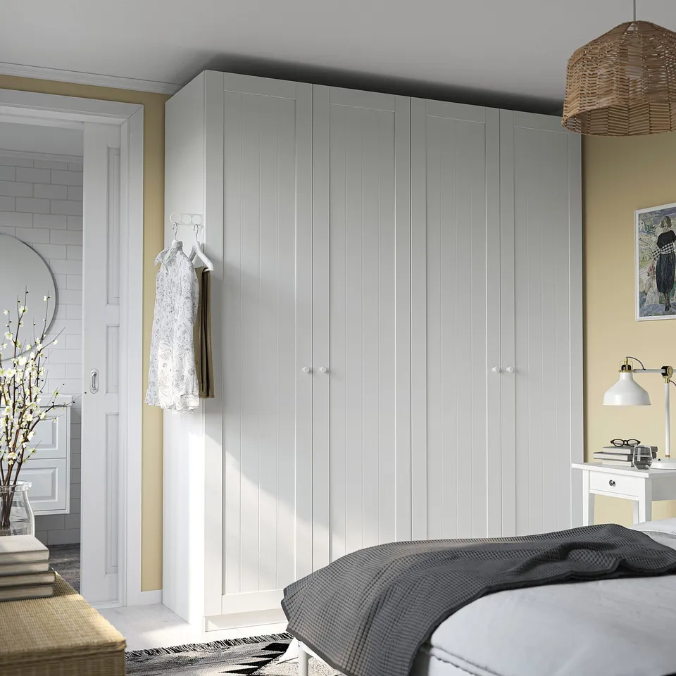 Minimalist bedroom with large white PAX wardrobe, hanging gown, bedside table with lamp, bed, and door to reflective bathroom.