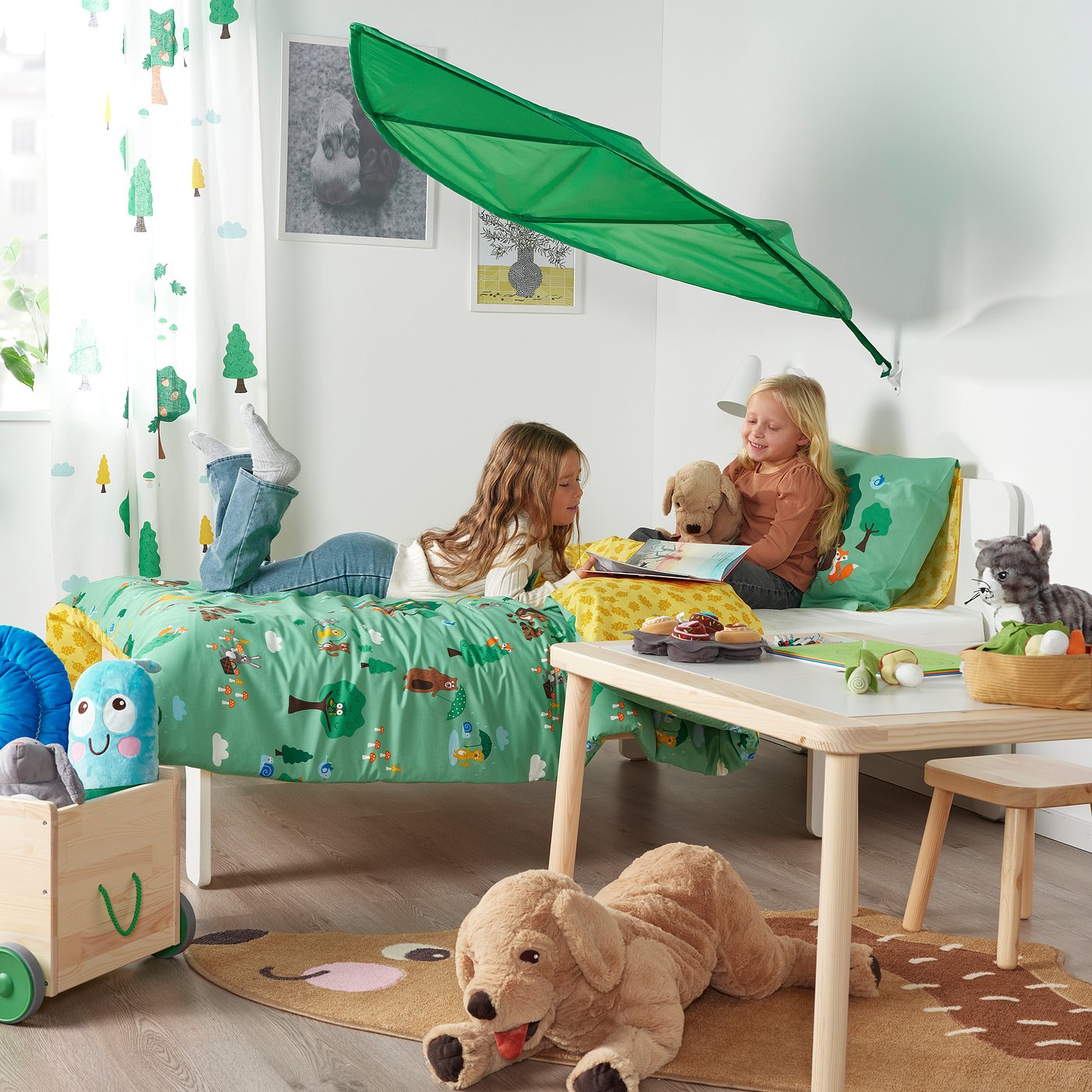 Two kids playing on a bed with a green leaf canopy; toys and blankets around.