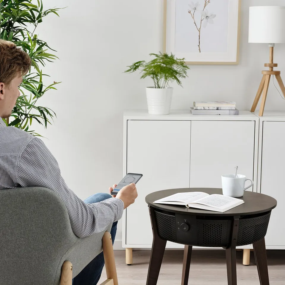 Person uses smartphone in chair. Table holds book, coffee, and STARKVIND air purifier.