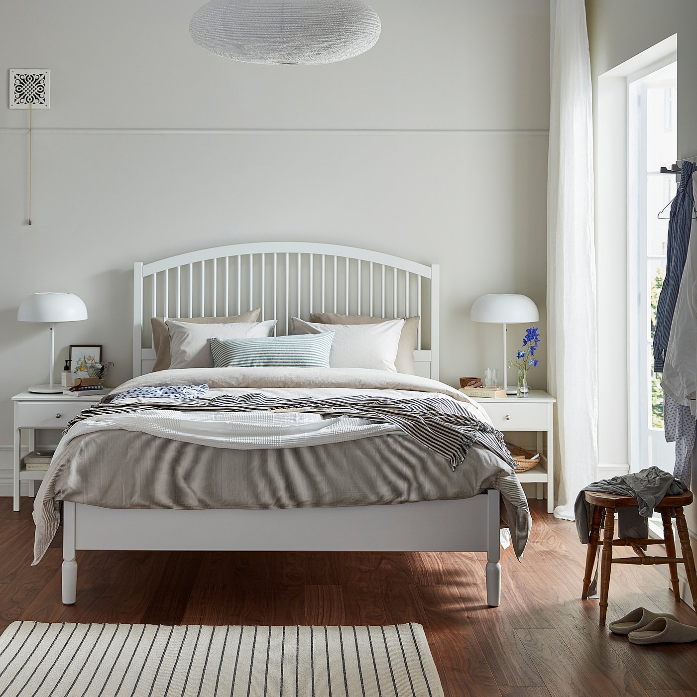Bright bedroom with TYSSEDAL white bed, striped linens, wooden flooring, two nightstands with lamps, and a large window with sheer curtains.