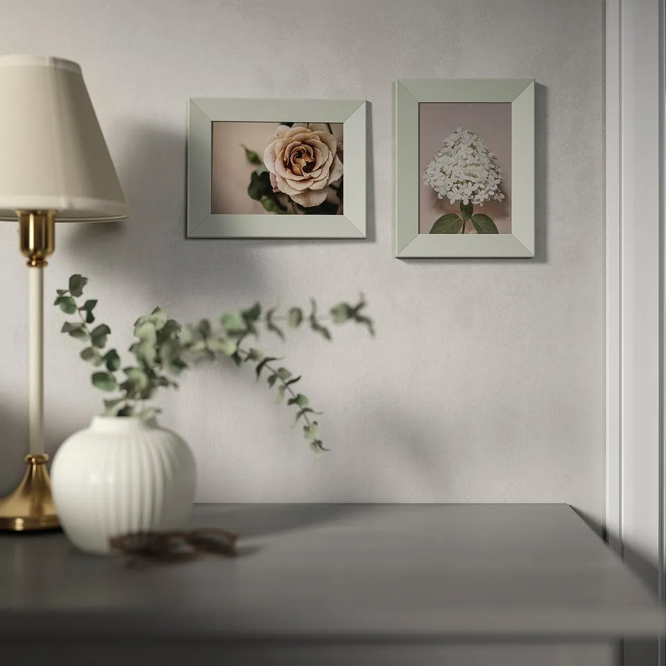 Two greenish frames hang on a wall, displaying close-up floral photos. Below them, a desk holds a lamp and a vase with eucalyptus.