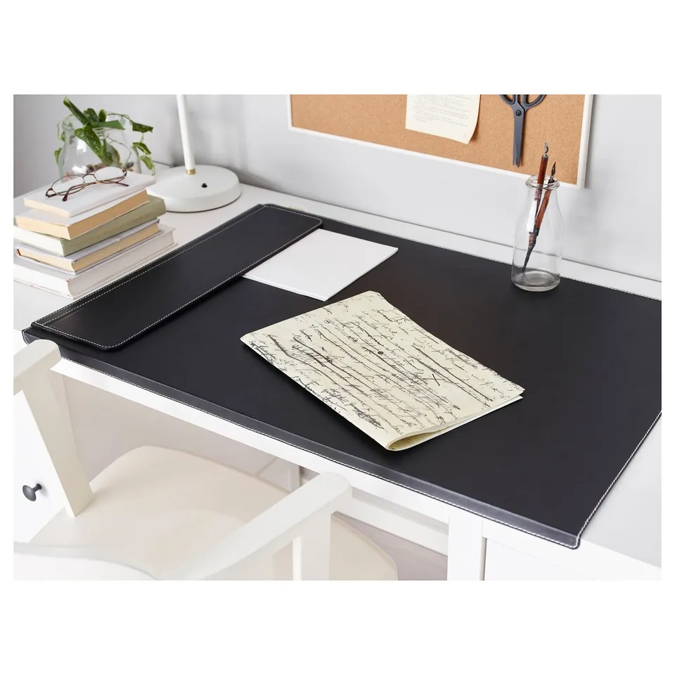 Black desk pad on white desk, with notepad, pen, books, lamp, and plant. Elegant, stain-resistant.