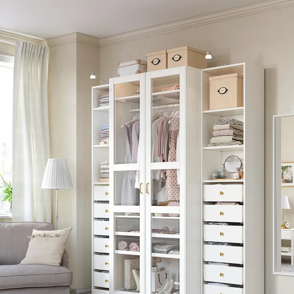 A modern, white PAX wardrobe with glass doors and drawers in a bedroom, featuring a grey sofa and rug.
