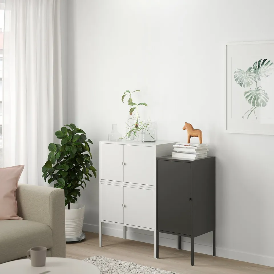 Modern living room with white curtains, grey sofa, LIXHULT white & black storage cabinets, green plants, wooden horse, and tropical leaf art.