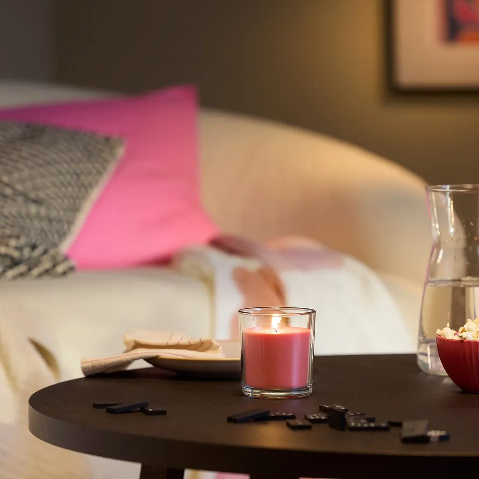 Pink LOTSFÅGEL candle, snacks, and games on dark table, cosy pink bed in background.