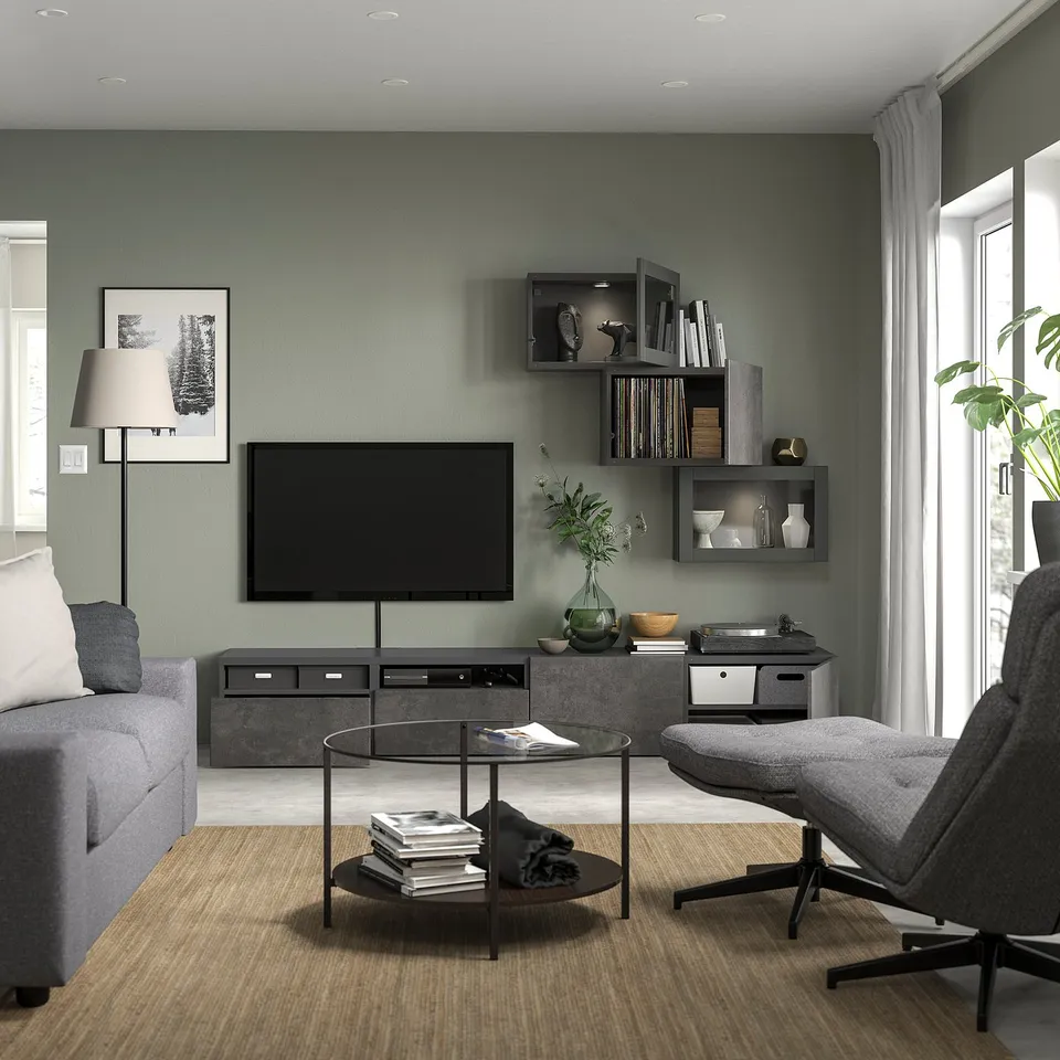Modern living room with grey walls, grey sofa, large tv, glass round coffee table with books, grey armchair, plant, and built-in shelves.