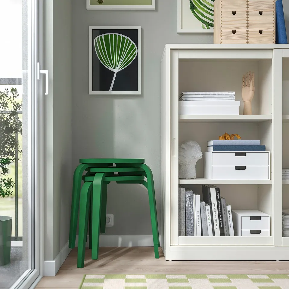 Modern room with white bookshelf and green stools. Books, decor, and storage boxes on the shelf. Stackable stools with three legs for stability.