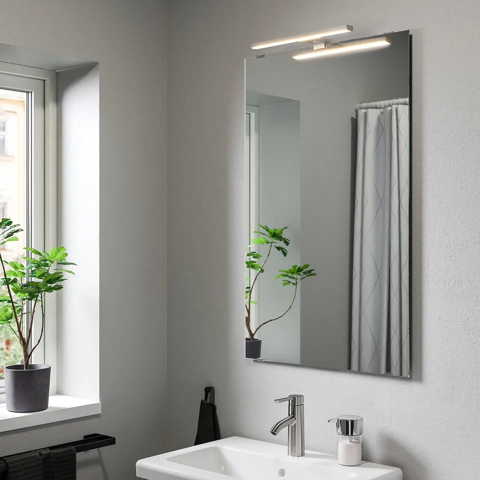 Modern grey bathroom with VÅTHULT mirror lamp, white sink, chrome fixtures, and plant on windowsill. Reflects curtain and room, with ample lighting.