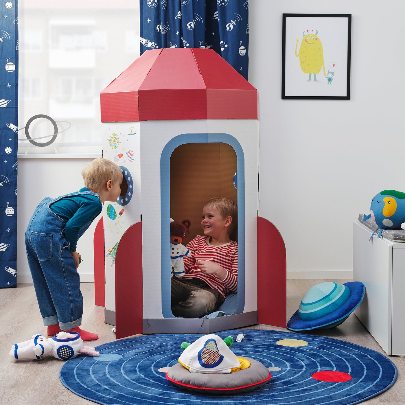 Kids play in diy rocket. Boy bends over, another sits with teddy. Astronaut toy on floor.